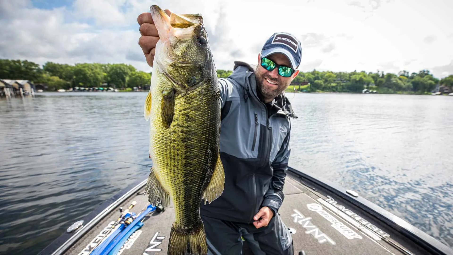 A huge largemouth bass being held up in front of a series of beautiful lakeside docks on Lake Minnetonka.